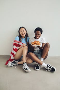Two diverse women celebrating Independence Day with a flag and burgers indoors.