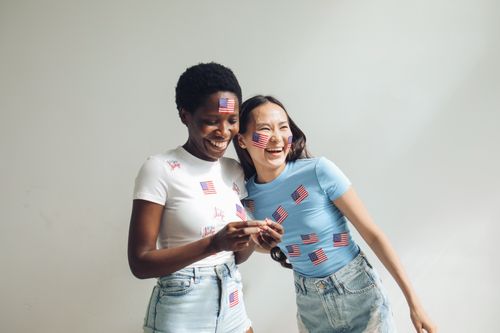 Free Two women joyfully celebrating with USA flags indoors, showcasing friendship and festive spirit. Stock Photo