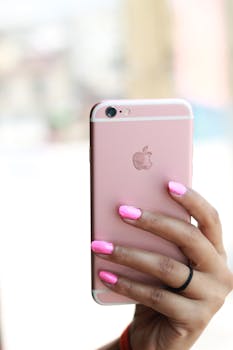 Vertical close-up of a hand with pink nails holding a smartphone, featuring a brand logo.