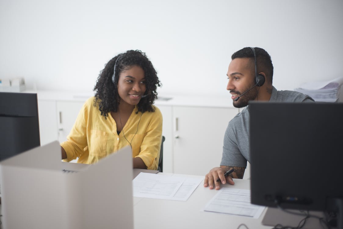 Free Woman in Yellow Dress Up Shirt with Headset Consulting to a Man Beside Her at Work Stock Photo