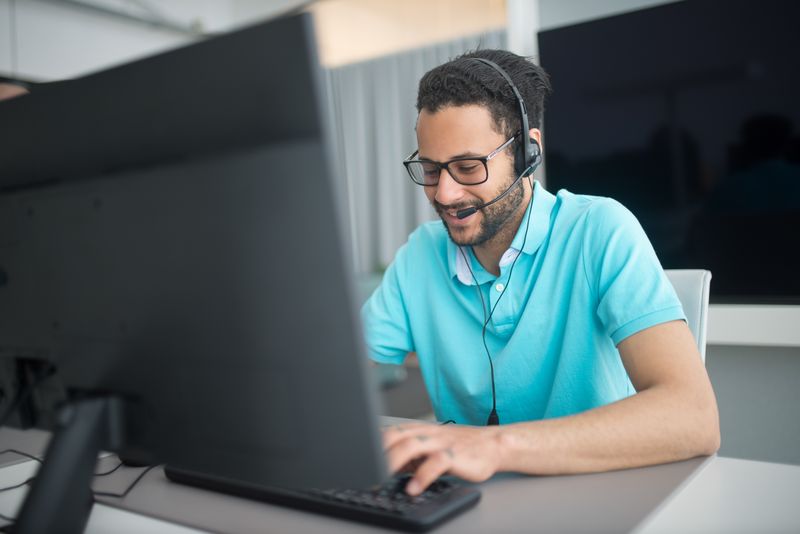 A man wearing a telephone headset typing on a laptop, with a monitor turned away from the camera in the foreground.