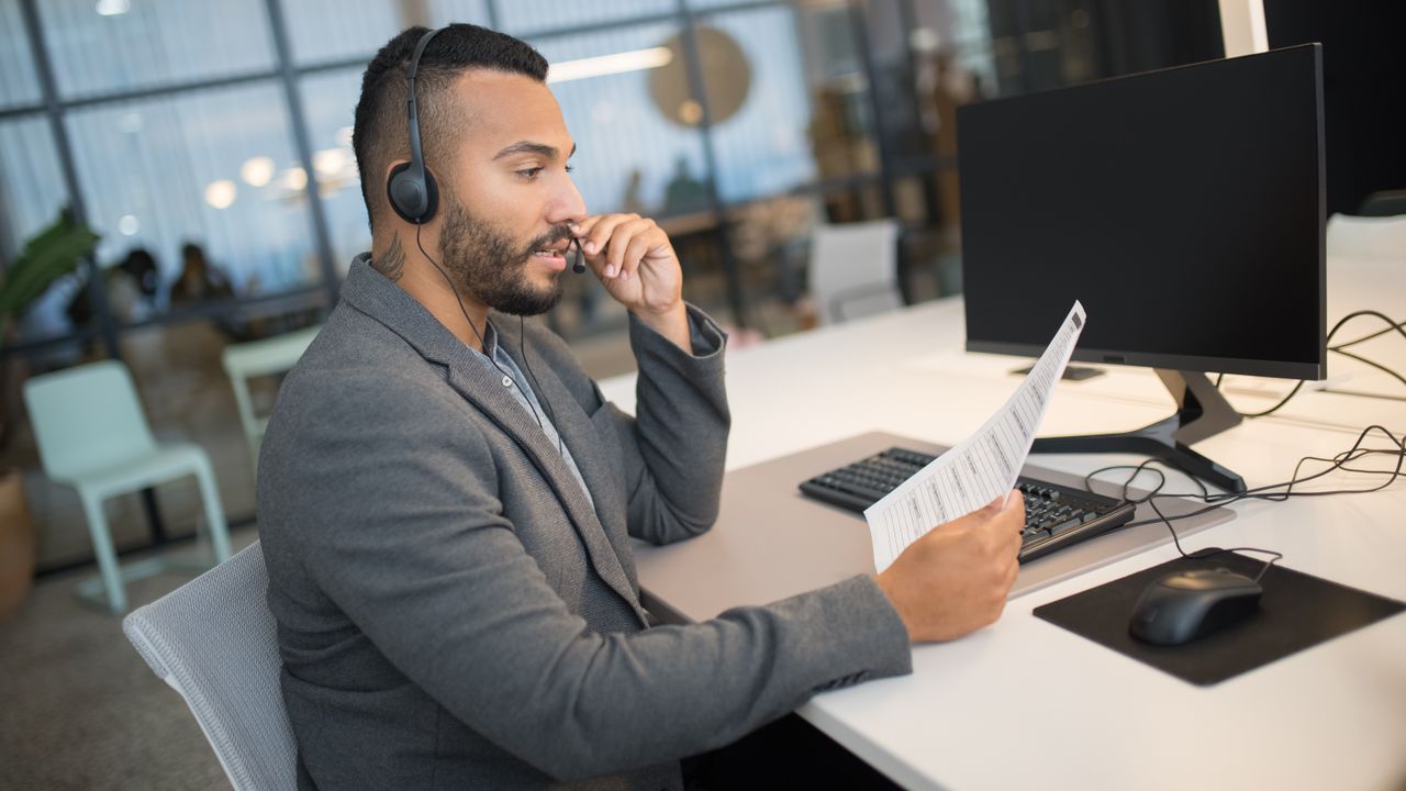 Young man in a call center, using a headset and reading documents at his office desk.