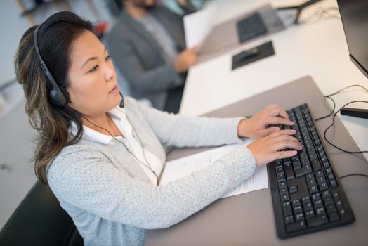 Asian woman working at a computer with headphones in a modern office setting.