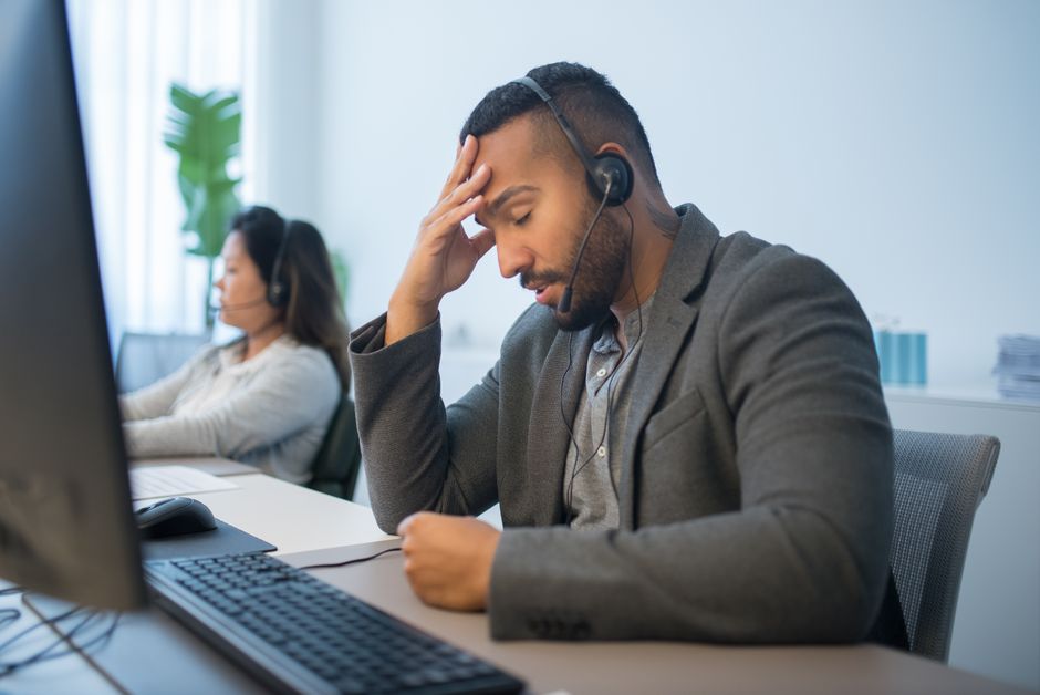 A tired call center employee with a headset looks stressed at his desk in a modern office.