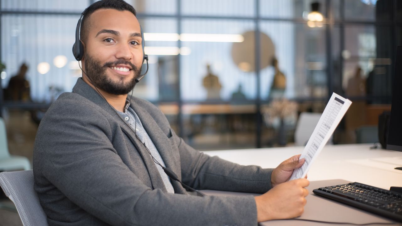 Smiling man wearing a headset, holding a document, sitting at an office desk.