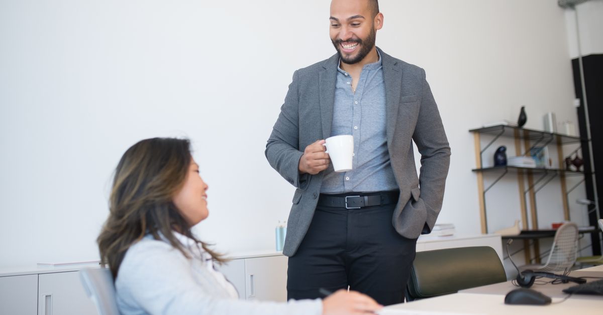 Two business professionals smiling and talking during a coffee break in a modern office setting.