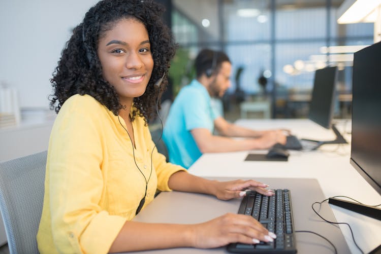 A Woman In Yellow Long Sleeves Wearing Headphones Sitting In Front Of A Computer While Smiling At The Camera