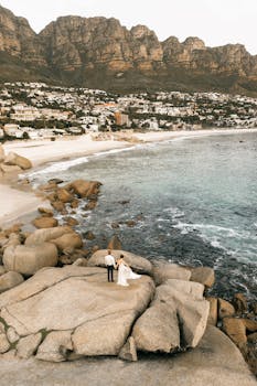 Aerial view of a newlywed couple on a rocky seaside cliff with mountain and ocean backdrop.