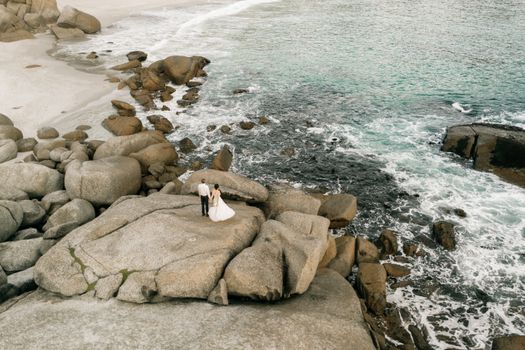 Aerial shot of a wedding couple on a rocky coastline by the ocean, capturing a romantic moment.