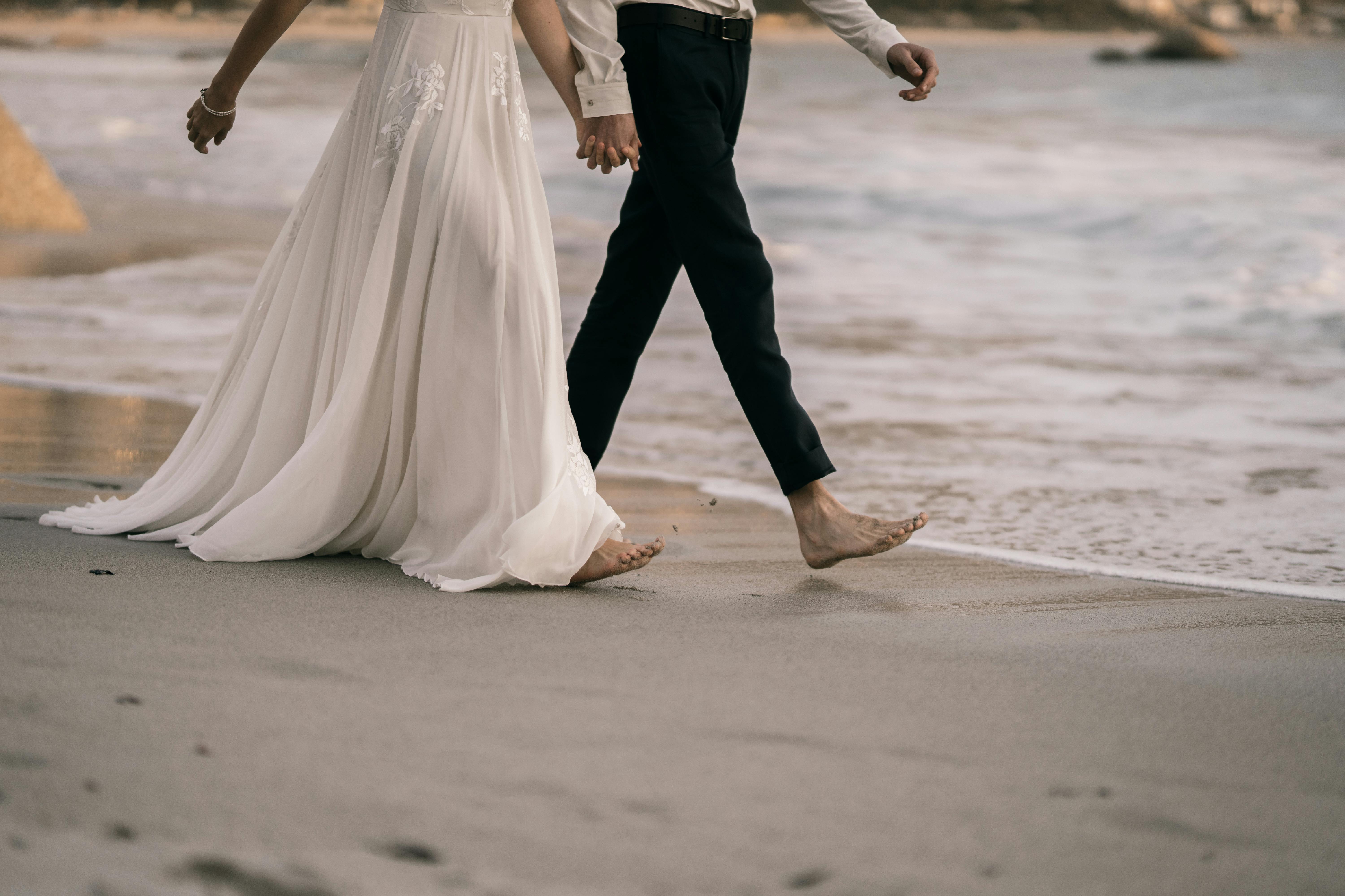 man and woman walking barefooted on seashore holding hands