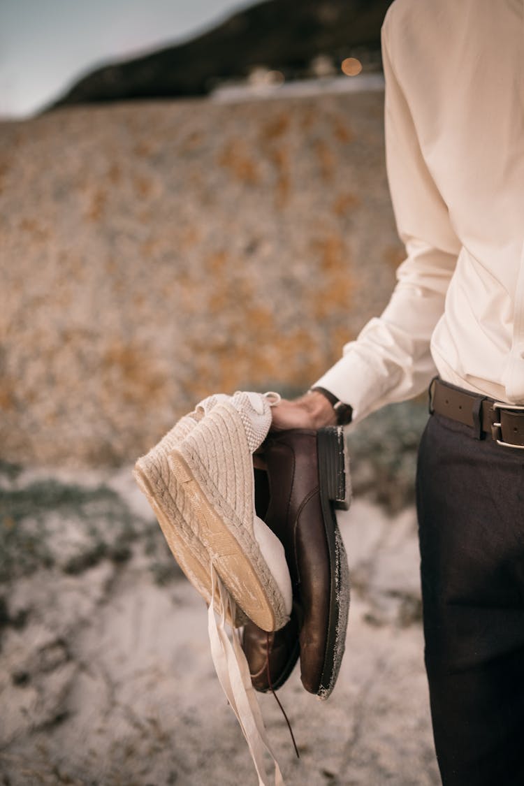 A Person Holding White Sandals And Brown Leather Shoes