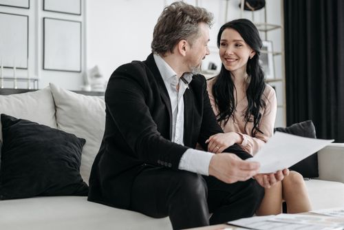 Free Couple smiling while sitting on a couch reviewing documents indoors. Stock Photo