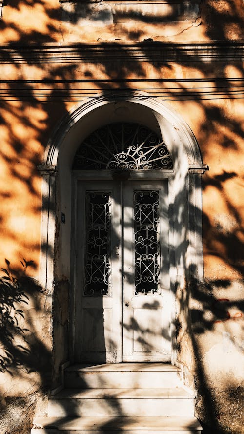 White door with arched passage of aged residential house with steps located on street with shadow of tree and bright sunlight in town