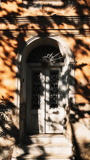 White door with arched passage of aged residential house with steps located on street with shadow of tree and bright sunlight in town