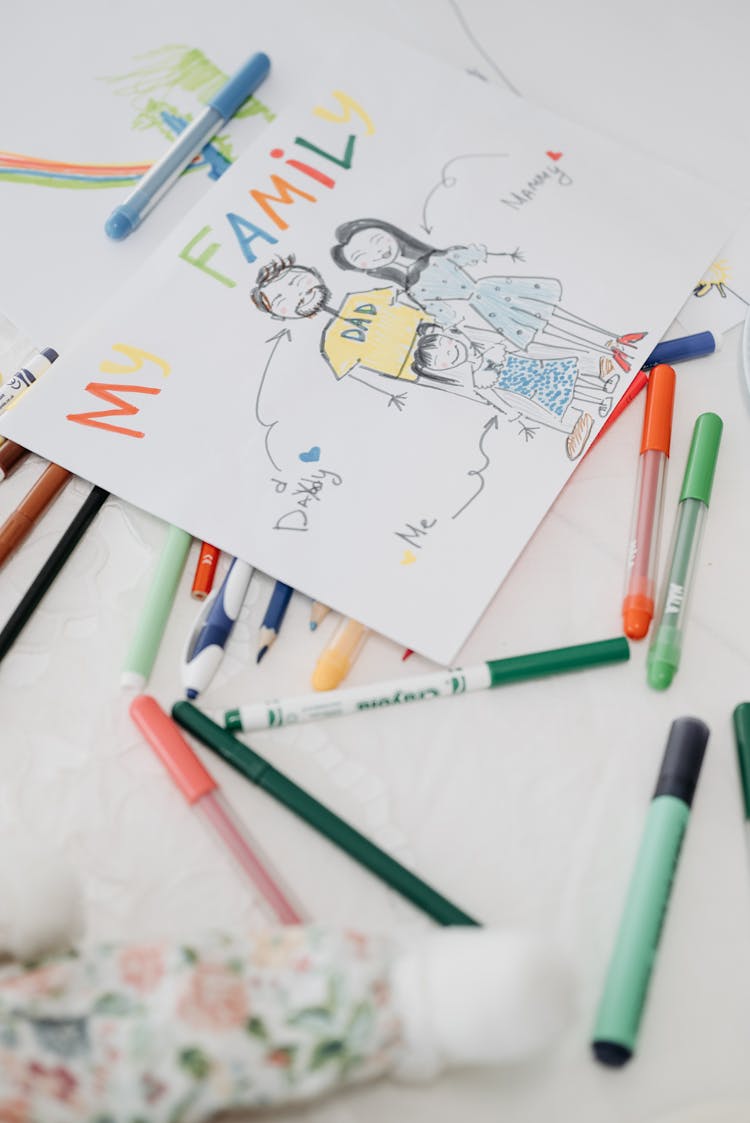 Childs Drawing Of A Family And Colorful Pens Lying On The Table 