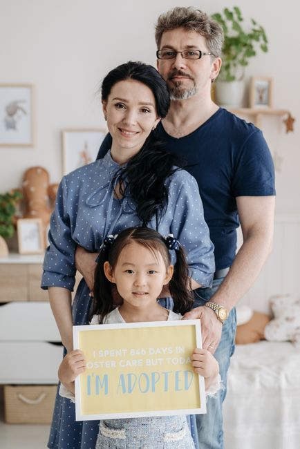 Happy family celebrating adoption with their smiling daughter holding an 'I'm adopted' sign indoors.