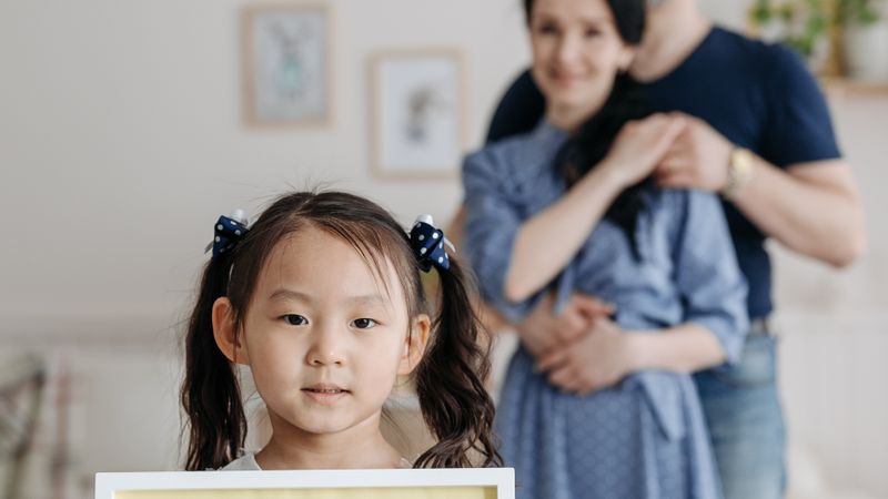 An adopted Asian girl holds a sign with her supportive parents in the background, symbolizing family and love.