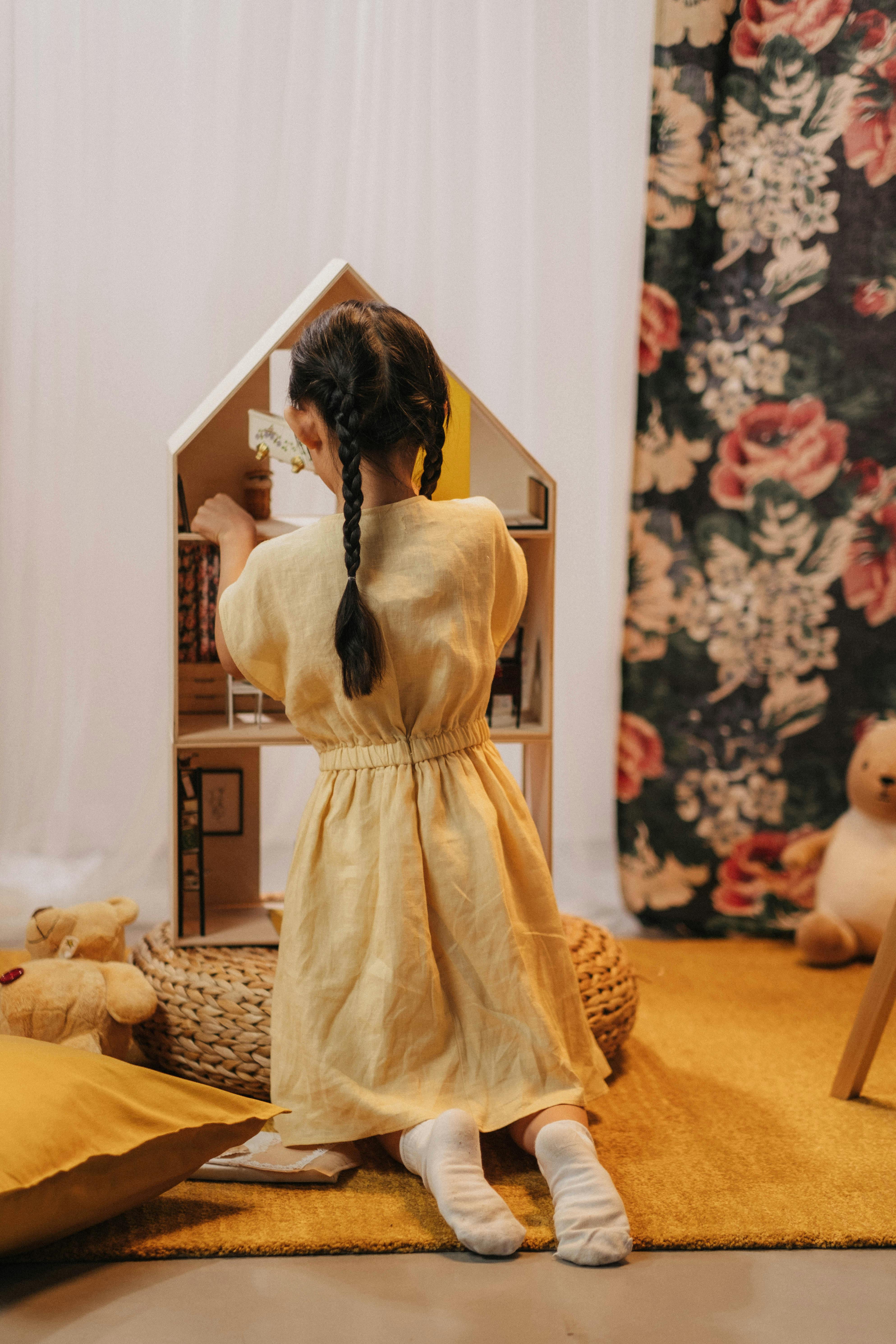 Free A young girl kneels while playing with a dollhouse, creating a playful childhood moment indoors. Stock Photo