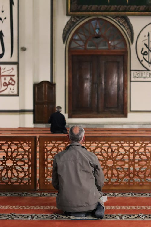 Free Back view of two Muslim men in mosque sitting on knees on floor covered with red carpet with ornament while praying Stock Photo