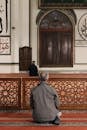 Back view of two Muslim men in mosque sitting on knees on floor covered with red carpet with ornament while praying