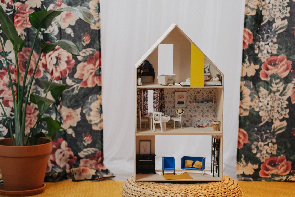 Finished miniature house displayed on a woven stool beside a houseplant, with tiny furniture on three floors
