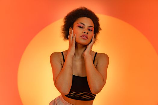 A stylish woman with afro hair poses against a vibrant orange background, showcasing fashion and beauty.