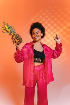 Cheerful woman in vibrant pink outfit holding pineapple against orange studio background.