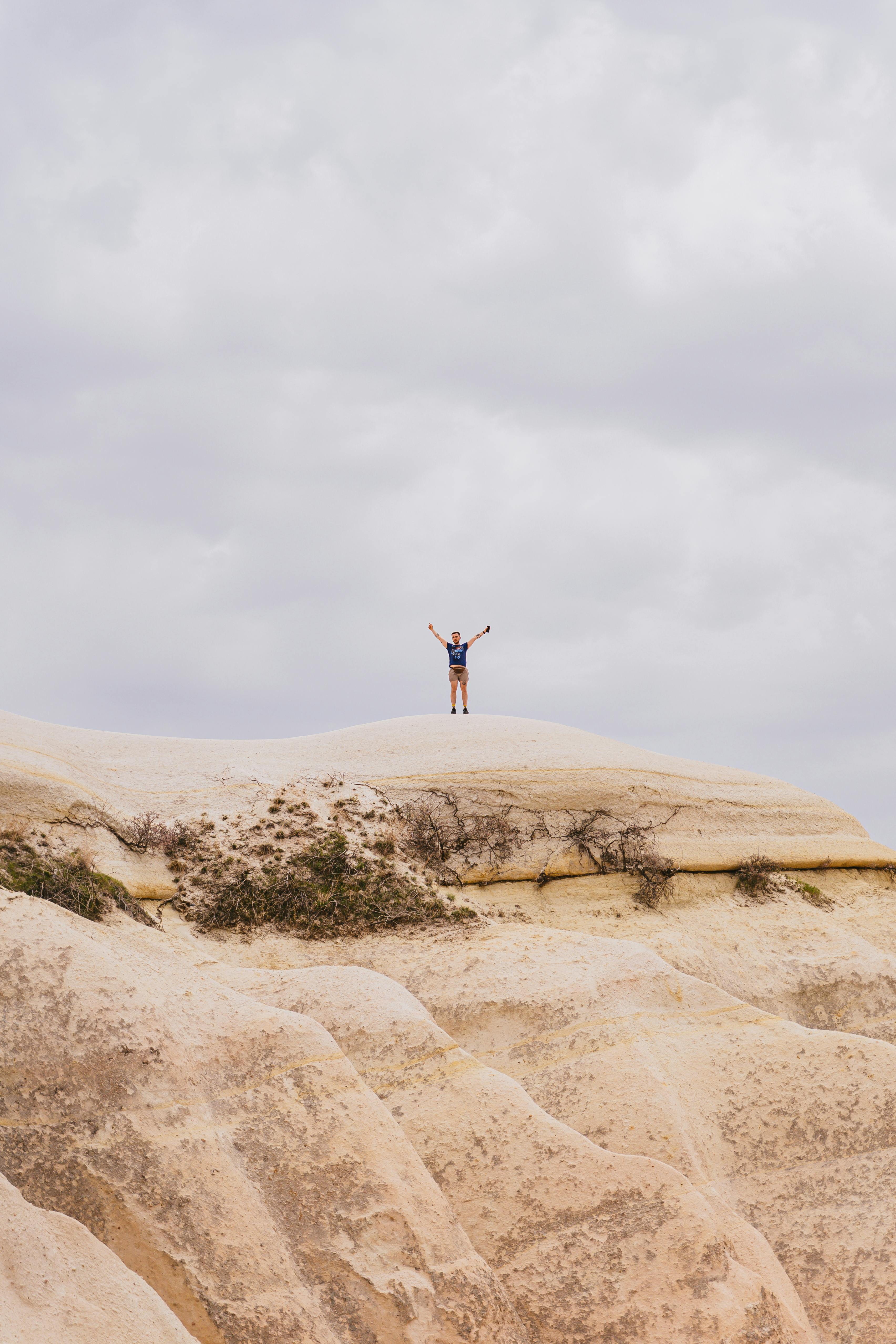 A man in shorts and blue shirt on a sandstone cliff, arms raised under cloudy sky.