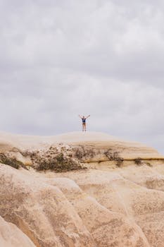 A man in shorts and blue shirt on a sandstone cliff, arms raised under cloudy sky.