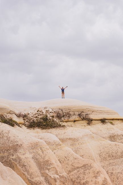 A man in shorts and blue shirt on a sandstone cliff, arms raised under cloudy sky.