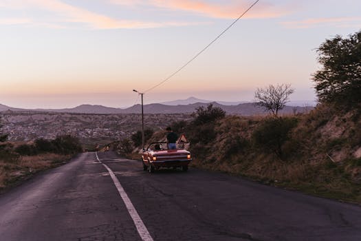 Red convertible with two people driving on a scenic mountain road at sunset.