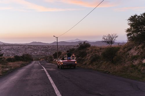 Free Red convertible with two people driving on a scenic mountain road at sunset. Stock Photo