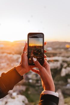 Hands holding a smartphone capturing a beautiful sunrise over a scenic landscape.