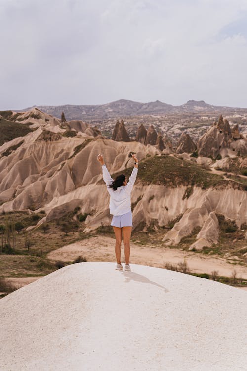 Free Woman in White Long Sleeve Shirt and Shorts Standing on Rock Formation Stock Photo