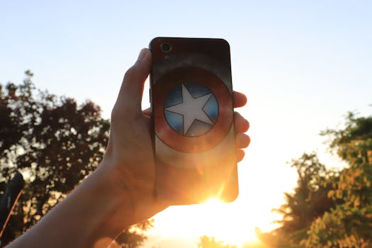 Hand holding a hero-themed smartphone case against a sunset backdrop with sun flare.