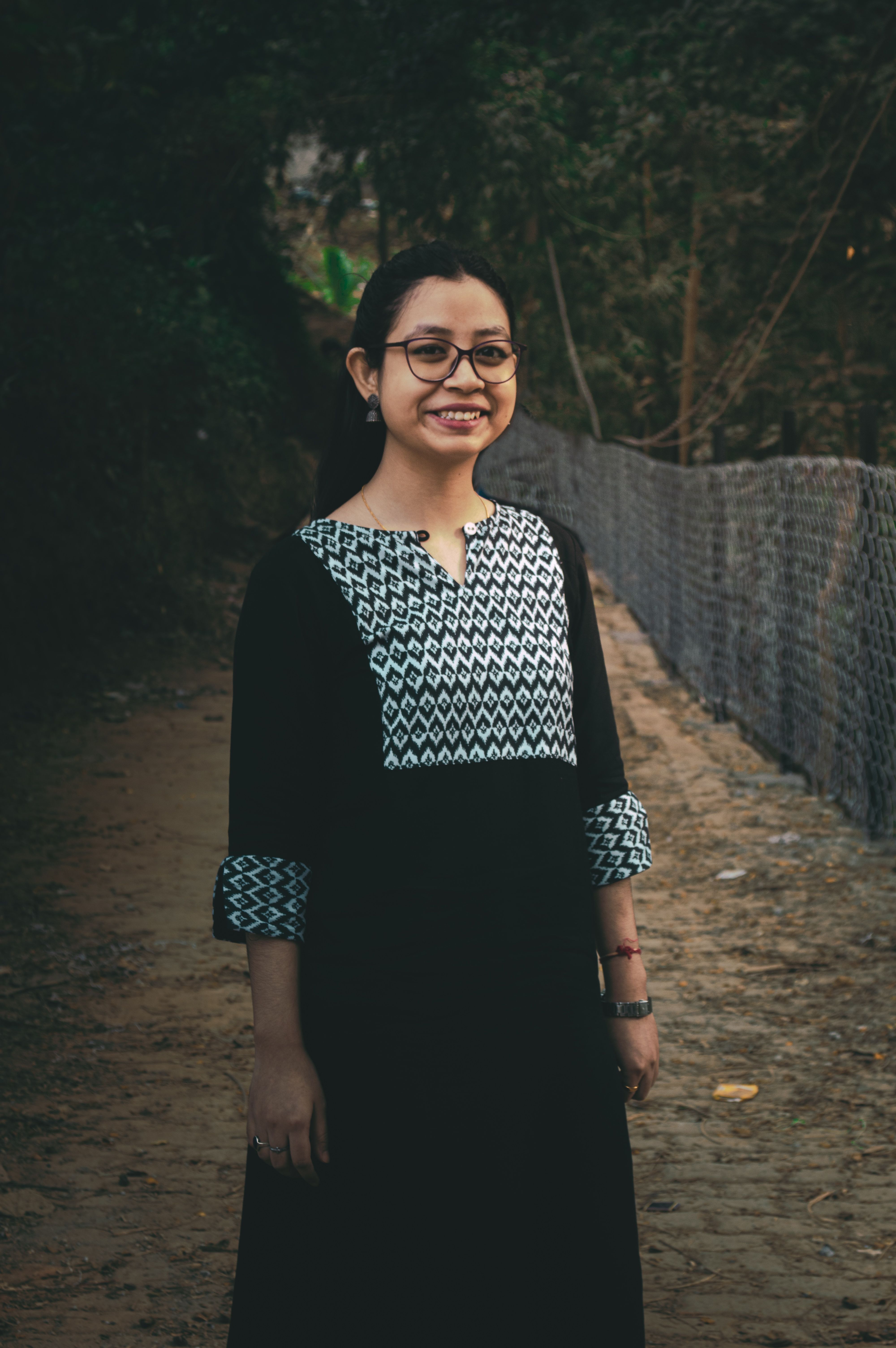 Portrait of a smiling woman wearing eyeglasses and a black dress, standing outdoors on a pathway.