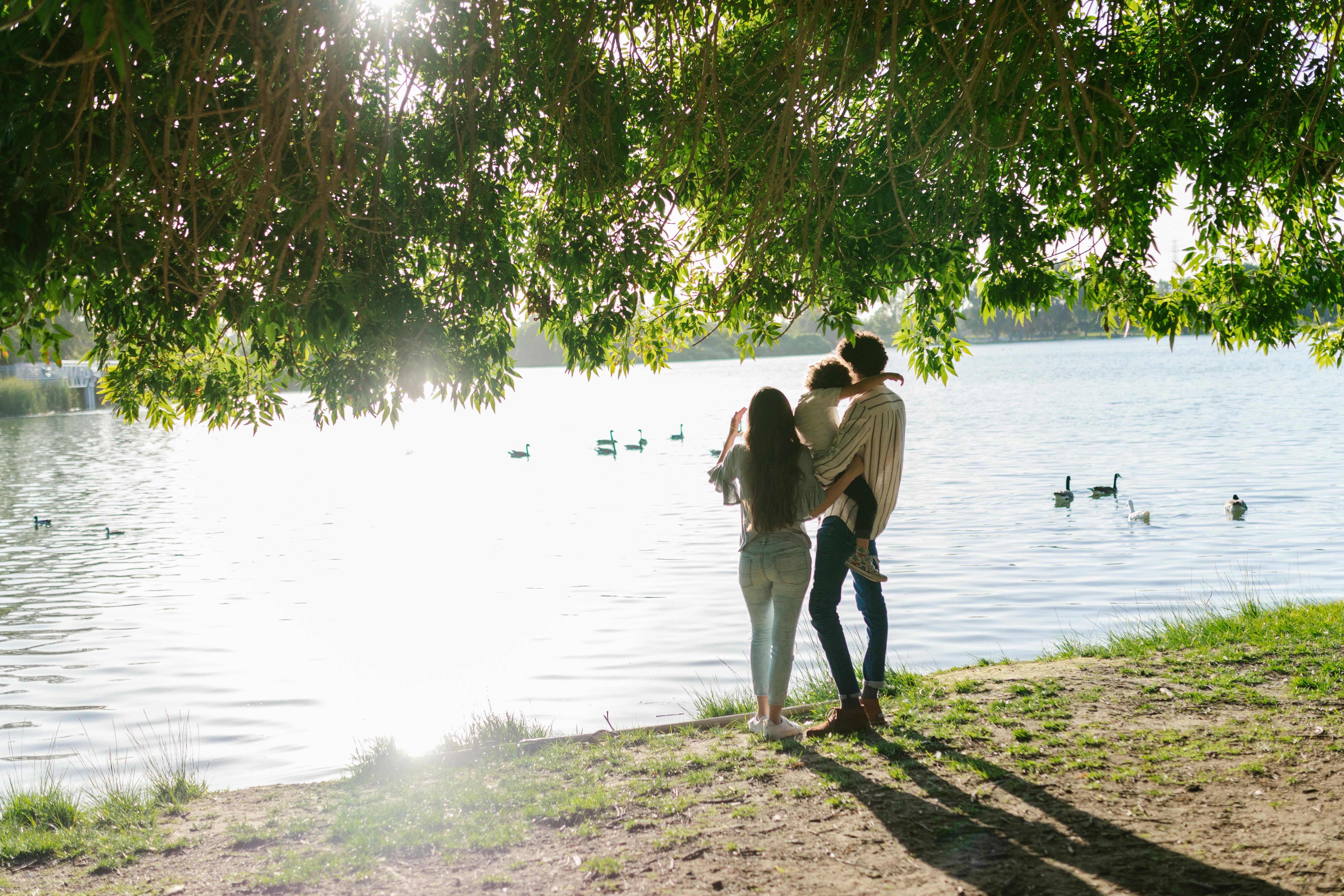 A young family enjoys a serene moment by the lake under a sunlit tree canopy.