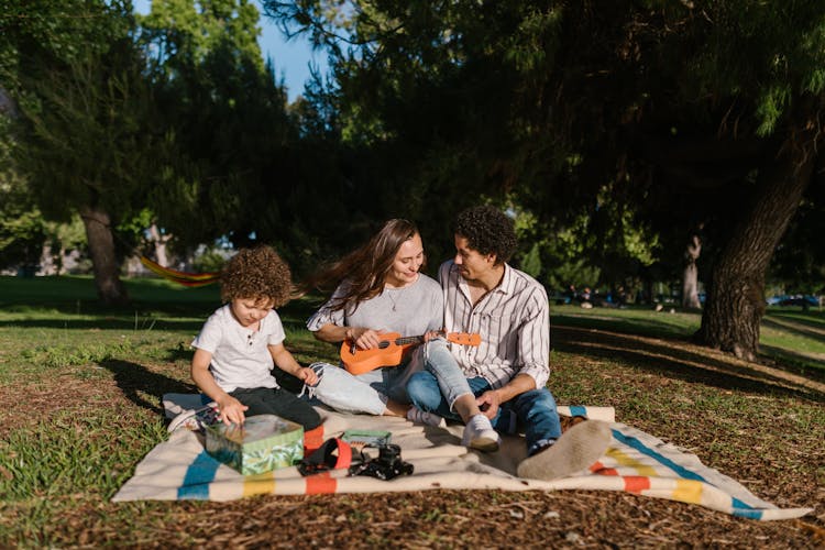 Family With A Little Son Having A Picnic In The Park And Woman Playing On Ukulele 