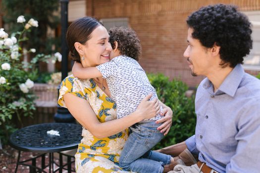 A joyful family moment captured outdoors as a child hugs their mother while the father looks on, all smiling.