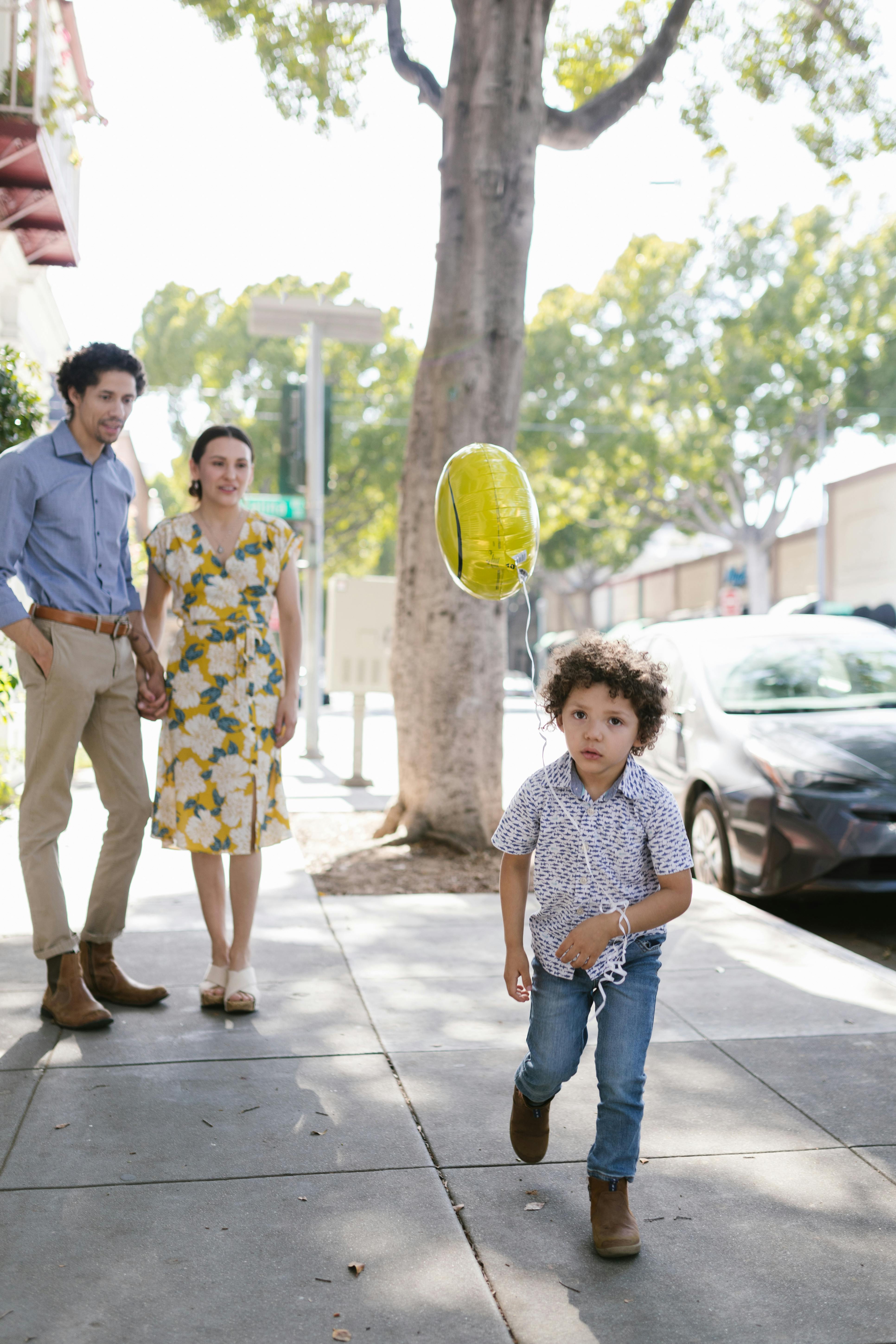 little boy with a balloon running along the sidewalk watched by his parents