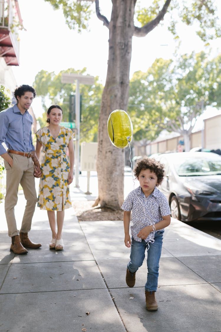 Little Boy With A Balloon Running Along The Sidewalk Watched By His Parents