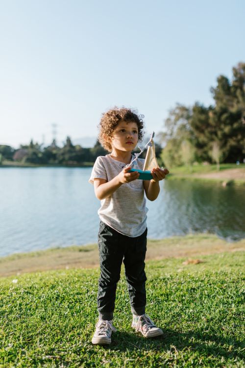 Free A young child with curly hair plays by the lake with a toy boat, capturing a moment of outdoor fun. Stock Photo