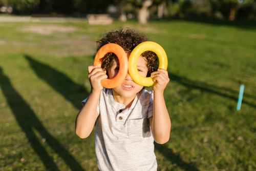 Free Smiling child holds colorful plastic rings up to their eyes, playing in a sunny park setting. Stock Photo