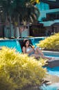 一名女子躺在泳池边喝着饮料，四周绿植环绕，还有几棵棕榈树