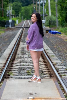 Woman in denim jacket smiling on railroad tracks, showcasing casual fashion outdoors.