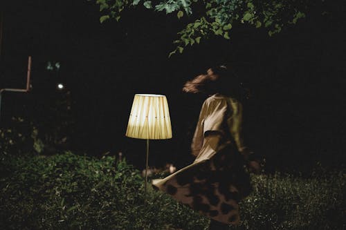 Back view of unrecognizable female in dress running in dark park with glowing floor lamp at night