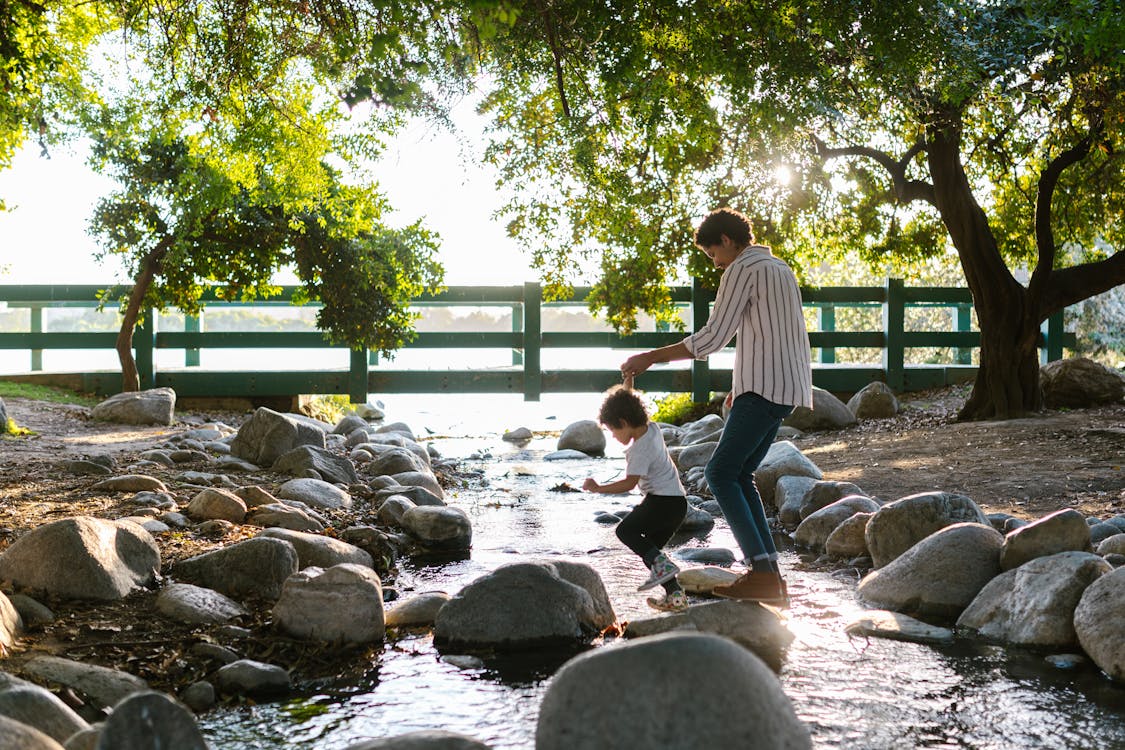 Woman Holding Hands with Her Little Son and Crossing a Stream by Walking on the Stones 