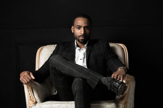 Stylish man in black suit sitting confidently on a chair against a dark background.
