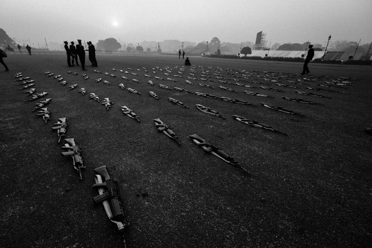 Grayscale Photo Of People Standing On A Field With Rows Of Rifles On Ground