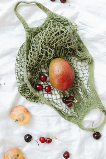Ripe mango and cherries arranged in a green reusable mesh bag on a white fabric surface.